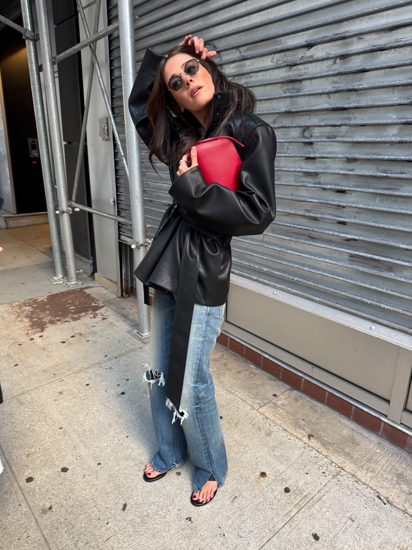 @ilanatorbiner wearing a black leather jacket, red bag, ripped split-leg jeans, and flip-flops.