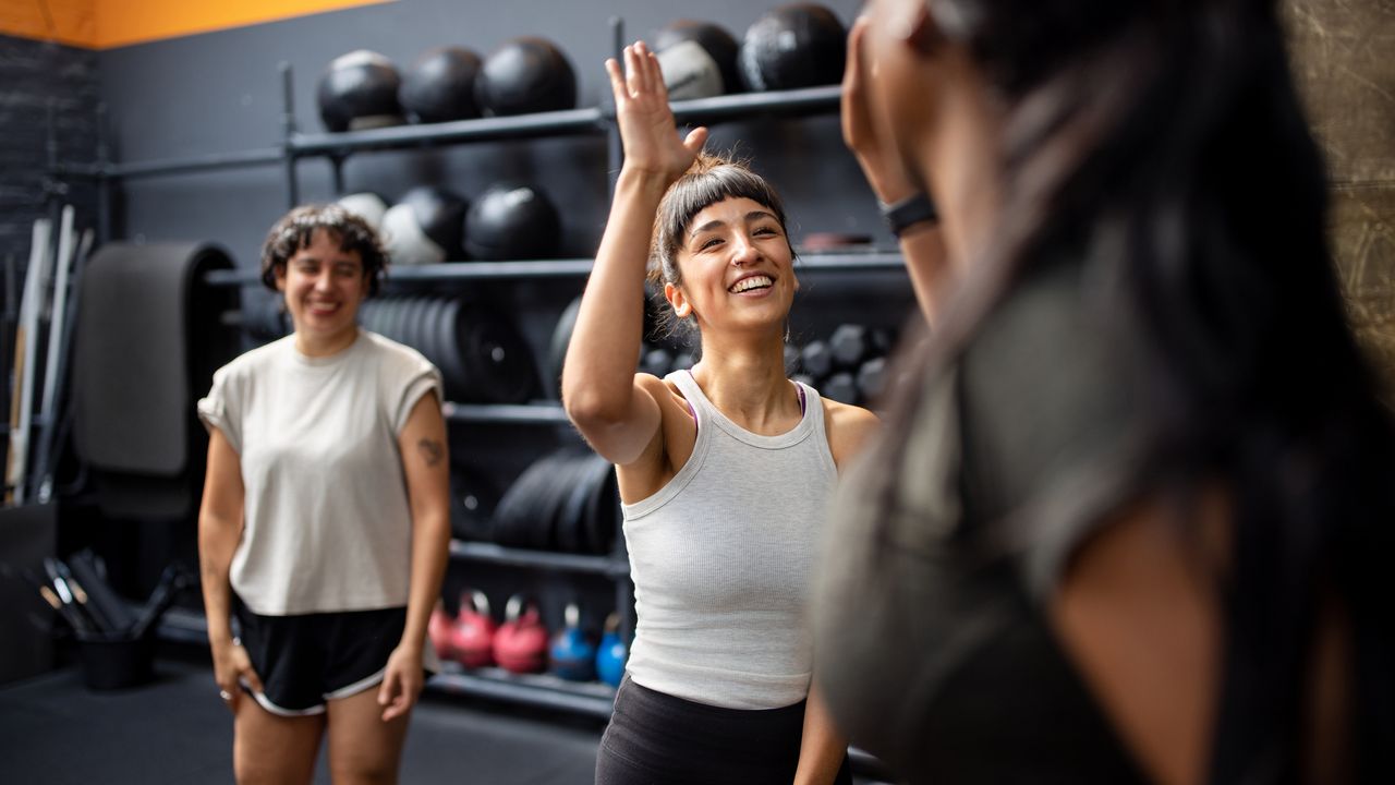 Two women high fiving in a gym while a third woman looks on from the background, smiling