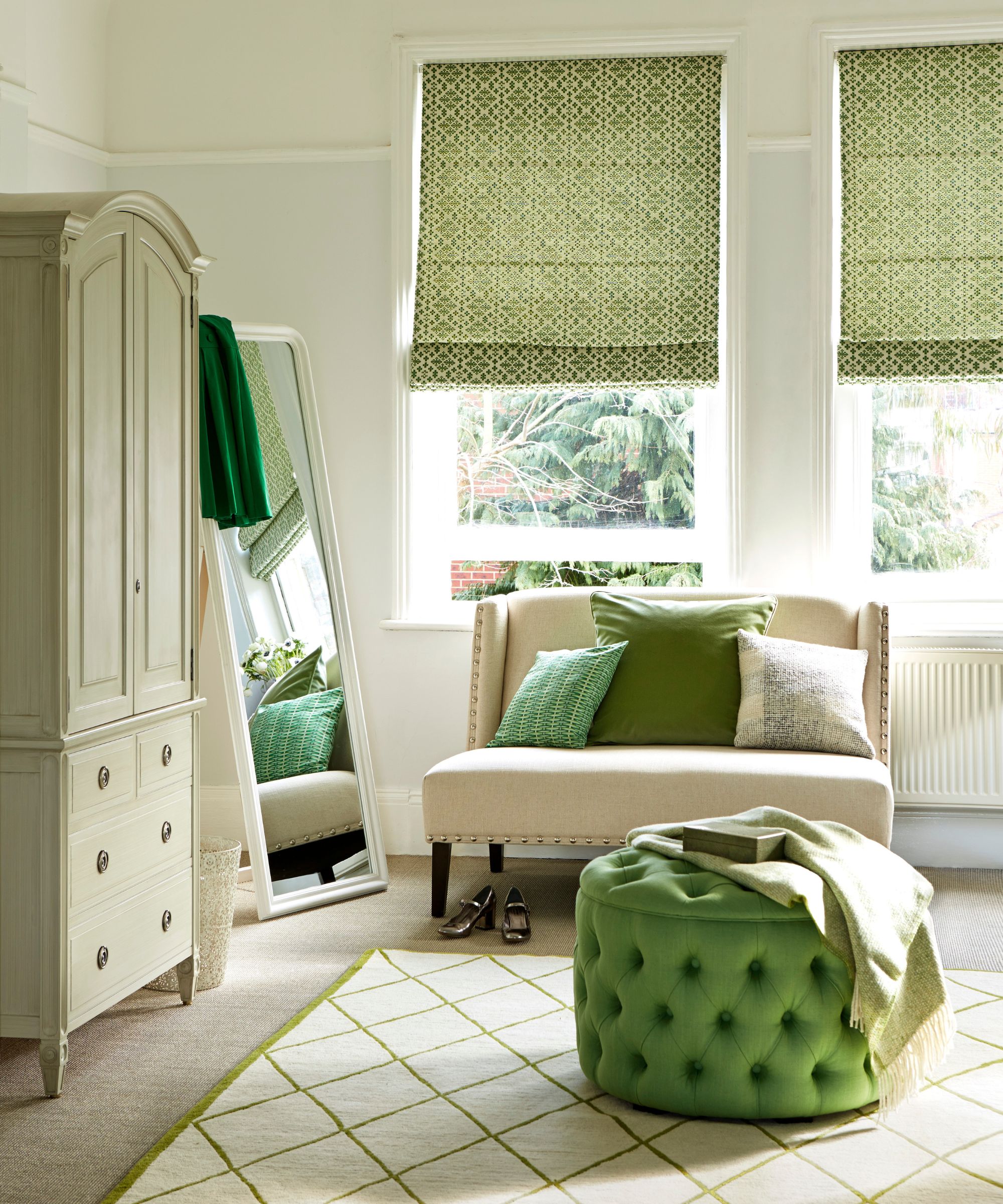 A white and green room with a large white closet, full-length mirror, white and green rug, green velvet pouffe, white couch with green cushions and two large windows with white and green blinds