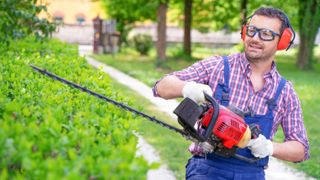 A man trimming a hedge while wearing safety goggles, gloves and ear protection