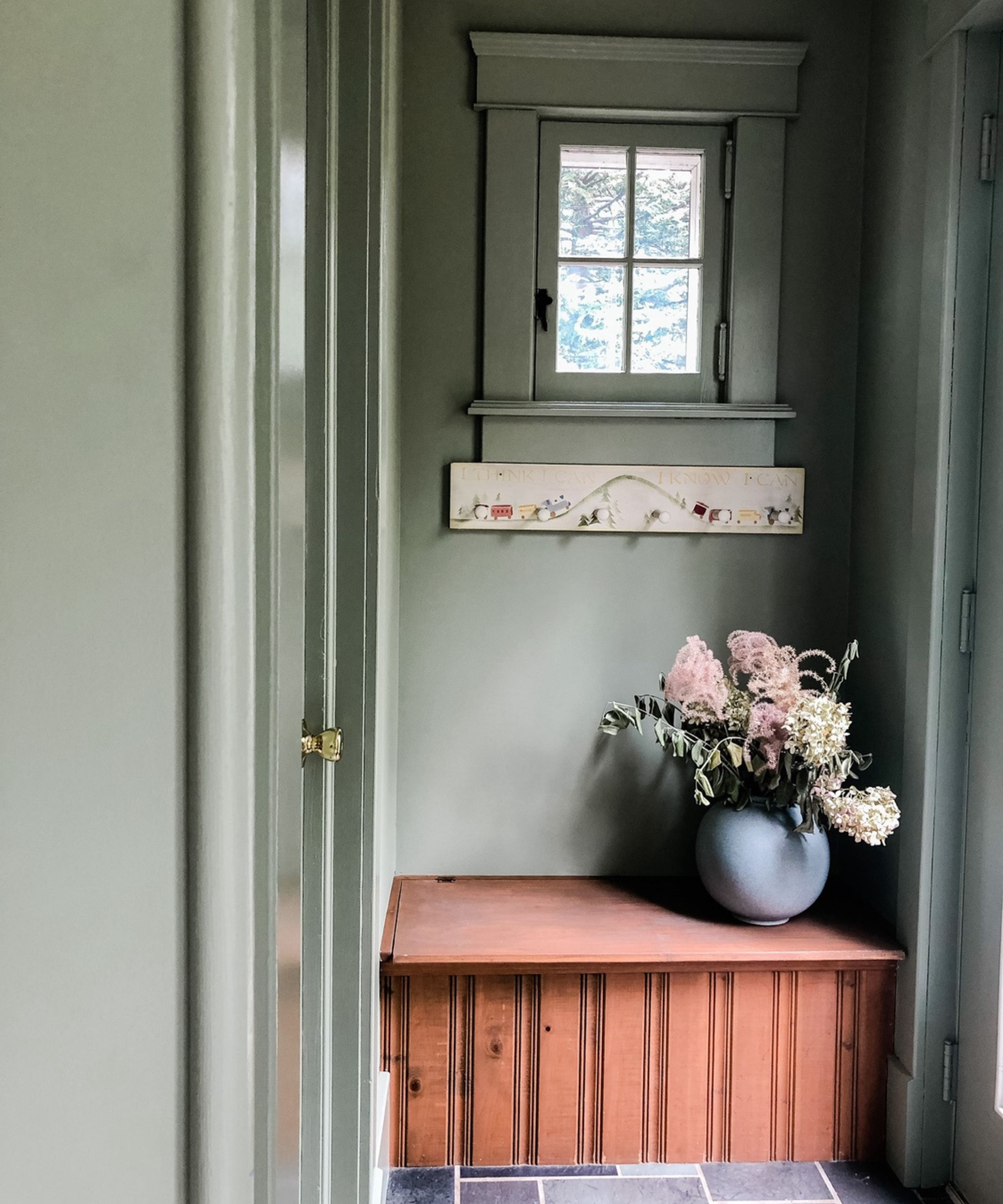 Mudroom with gray green painted walls, a built-in wooden bench, coat hooks and a small window and a vase of flowers