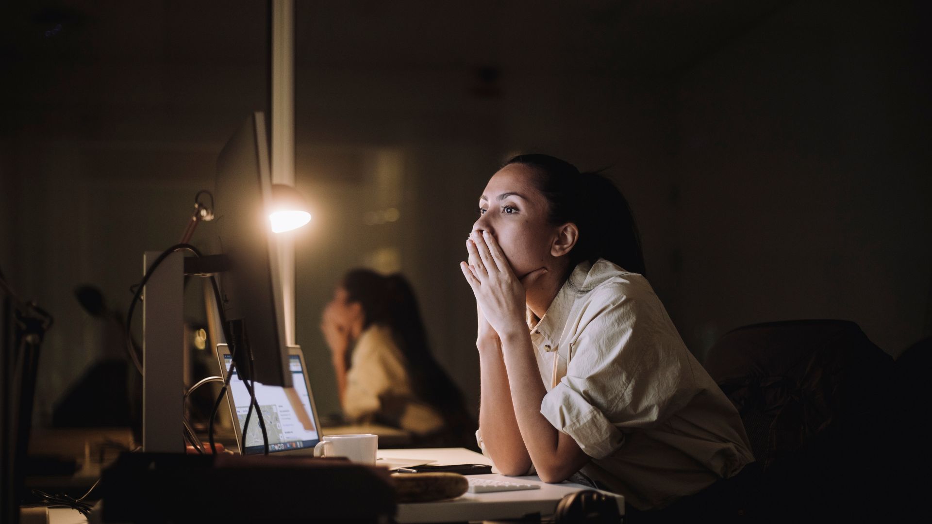 A woman sitting at her desk at work at night with her hands over her face, looking stressed.