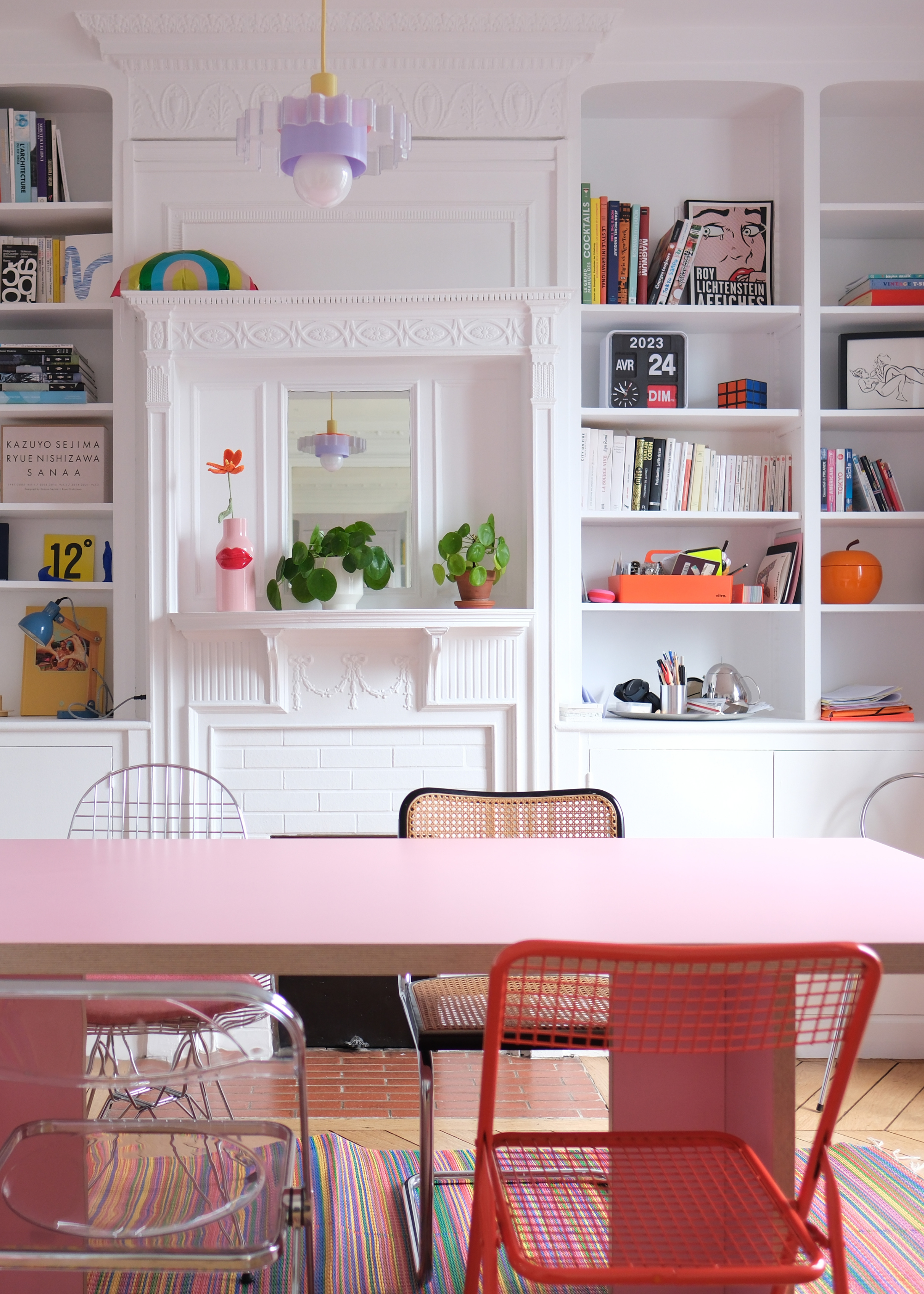 A white living room with in-built shelving featuring colorful books and knick knacks, a pink dining table with mismatched metal and rattan chairs, potted plants, and a pendant purple light