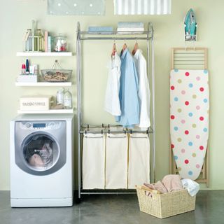 A laundry room with a Hotpoint washing machine, open shelving with washing powder in the vintage tin, linen water, washing caddy, sewing kit, clothes rail with clean shirts hanging, a traditional airer hanging from the ceiling, spotted ironing board, iron attached to the wall and a laundry basket with dirty towels