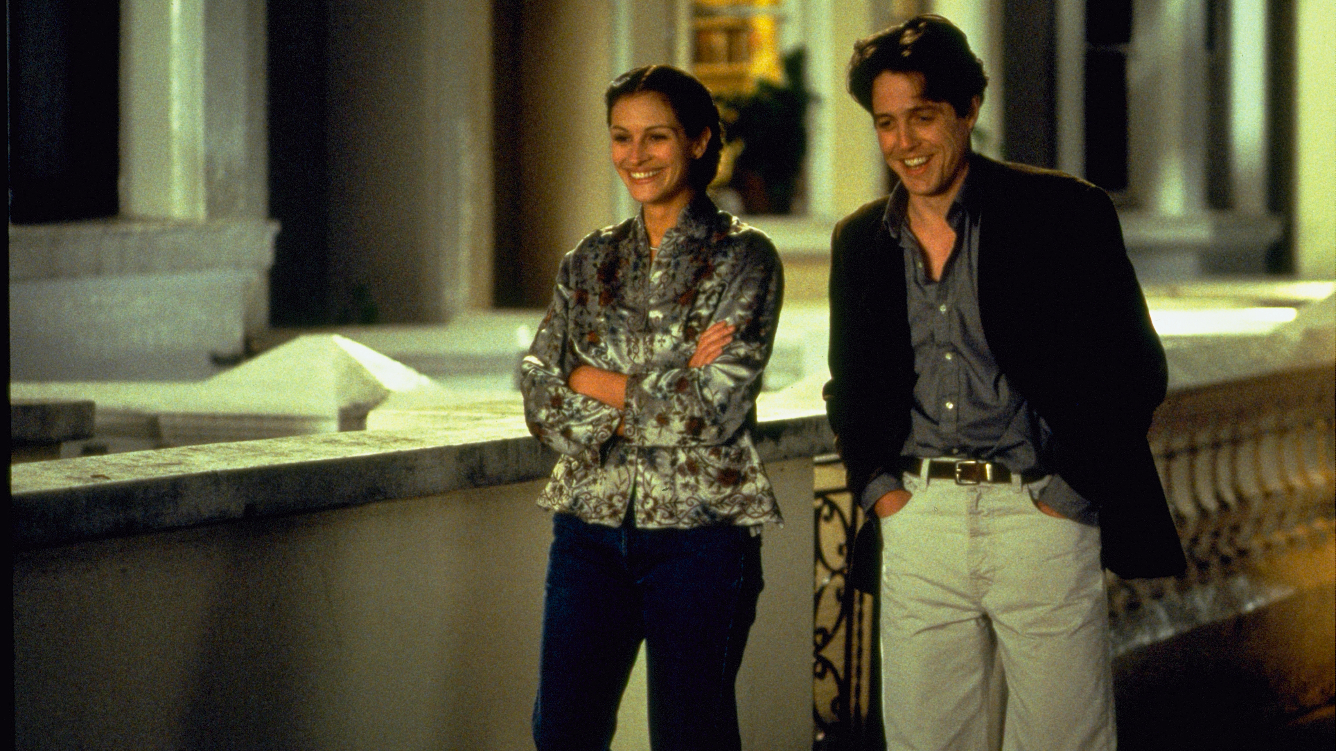 A still from the movie Notting Hill in which Hugh Grant plays Will Thaker and Julia Roberts plays Anna Scott, they’re both walking down a street in London smiling.