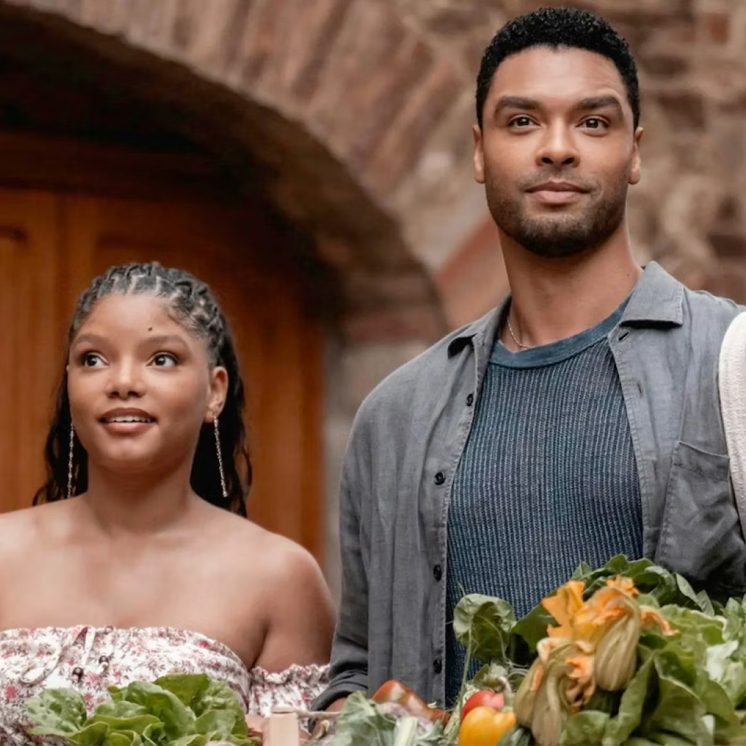 halle bailey and rege jean page smiling on a balcony with produce in front of them in you me and tuscany