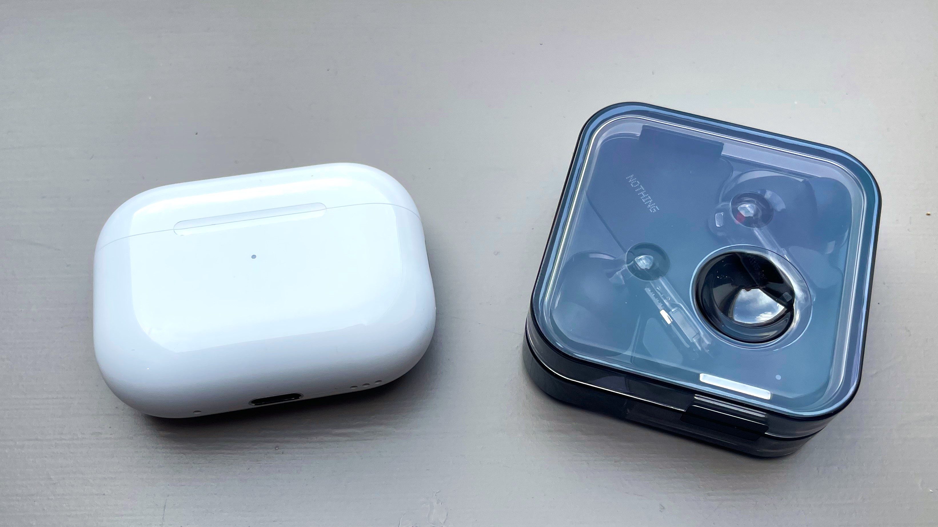 Apple AirPods Pro 2 vs Nothing Ear charging cases side by side on a gray painted surface