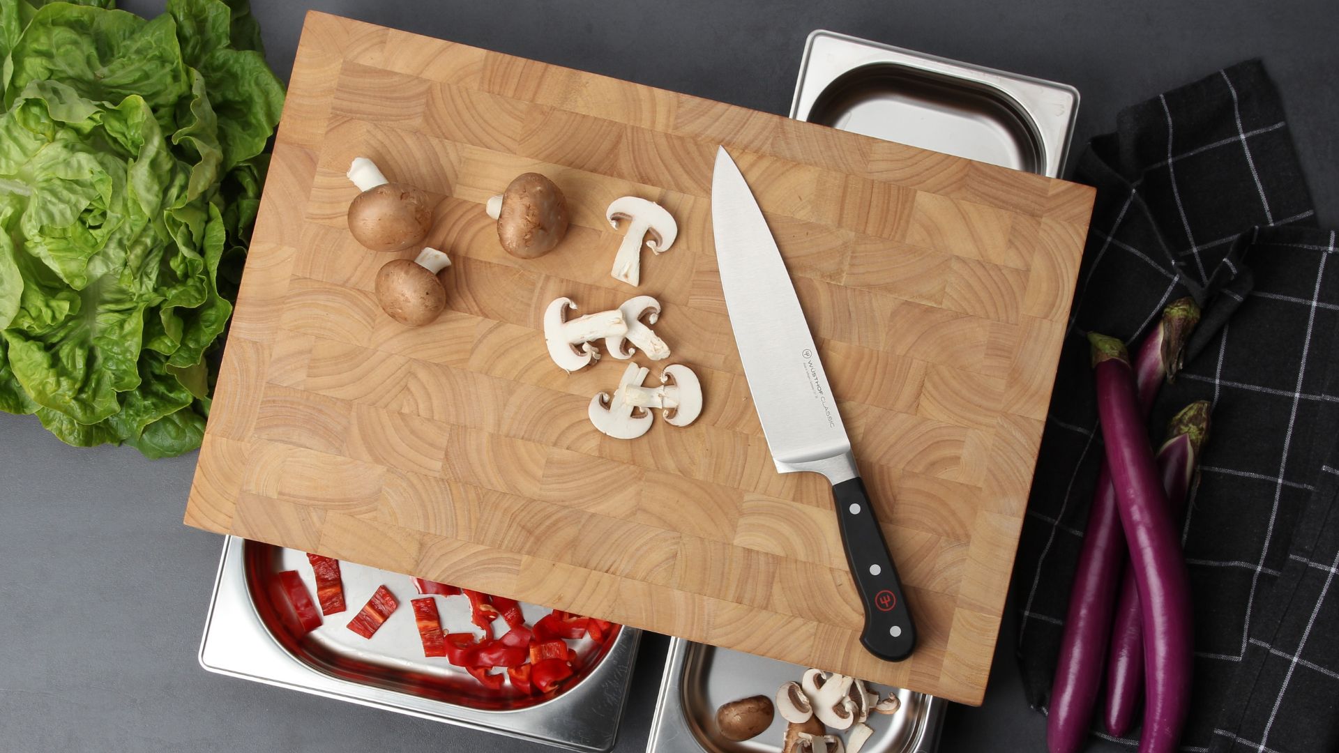 A wood cutting board with silver trays, chopped mushrooms, a head of lettuce, aubergine and a black washcloth