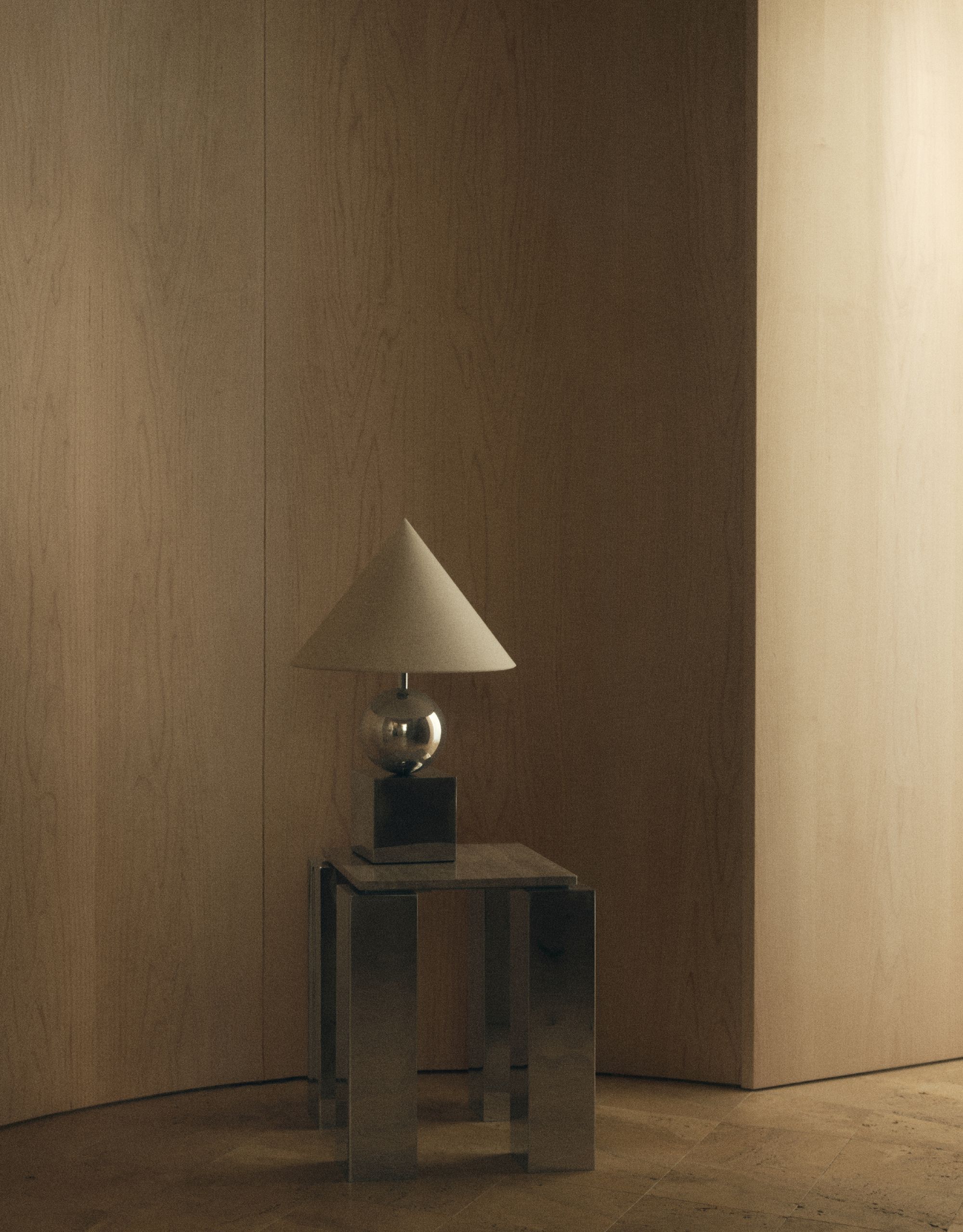 A modern chrome and travertine side table with a geometric chrome table lamp with a linen cone shade sits alone in front of a curved wooden wall on wooden flooring