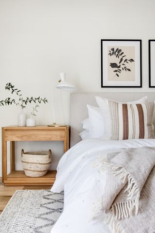 white bedroom with oak wood nightstand, artwork, white desk lamp, textured stone blanket, white bedding, stripe cushion