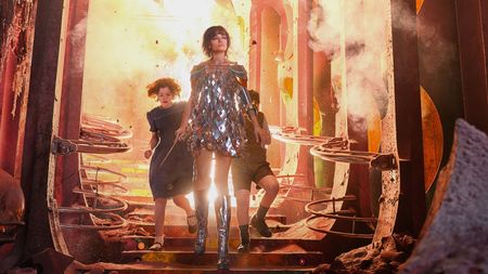 Jadis, in a mirrored minidress, shoulder armour and silver knee boots, strides down steps out of an explosion in a crumbling, rose-red palace, with Polly and Digory behind her