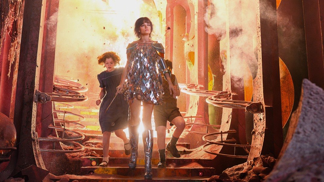 Jadis, in a mirrored minidress, shoulder armour and silver knee boots, strides down steps out of an explosion in a crumbling, rose-red palace, with Polly and Digory behind her