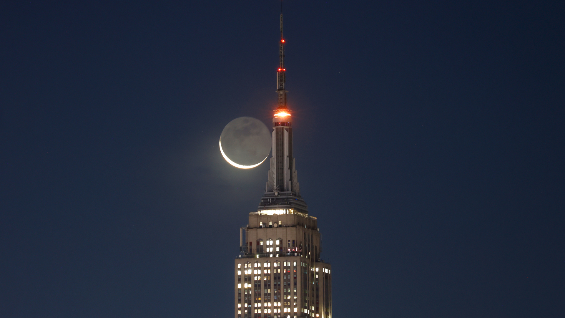 A thin crescent moon is shown next to the upper spire of a skyscraper in a dark sky. Its unlit surface is suffused with a soft glow, revealing the presence of lunar seas, or mare, on its surface.