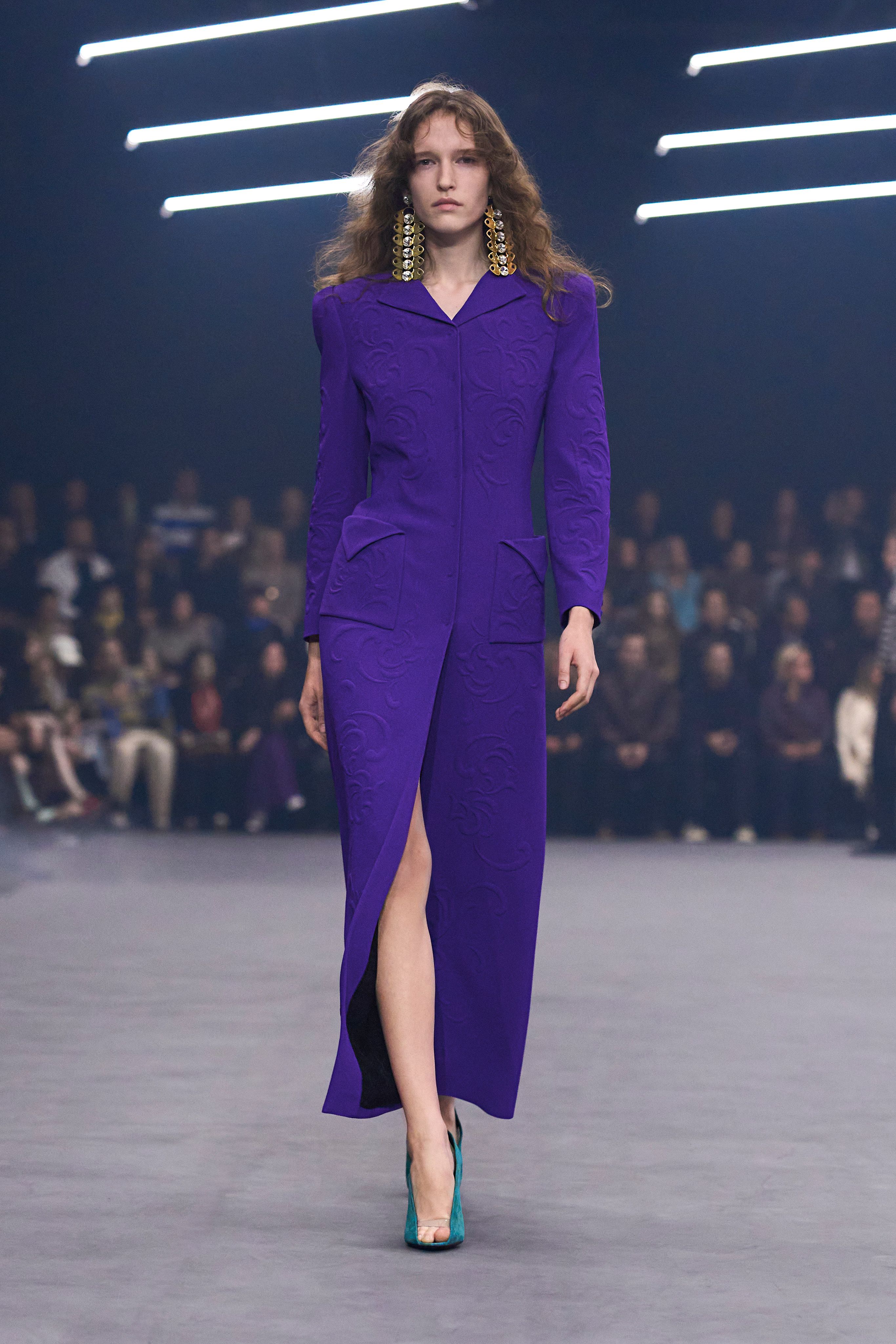 A Valentino model wearing a tailored purple coat styled as a dress, oversize art deco earrings, and a pair of cerulean peep toe heels.