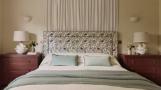 a bed with a floral headboard, two dressers on either side with lamps on top, green pillows, and a curtain behind the bed