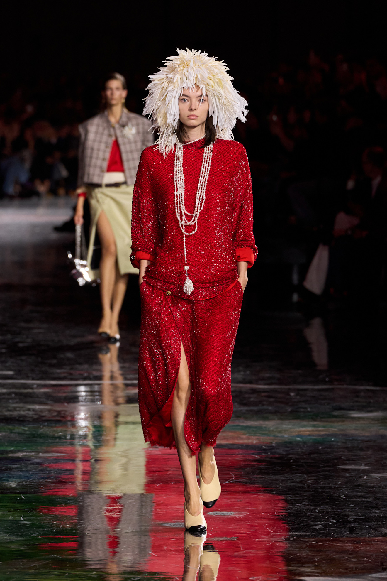 A Chanel model wearing a sparkly red knit shirt with a matching high-slit skirt, two-tone cap toe heels, a long white beaded tassel necklace, and a white feather headcap.