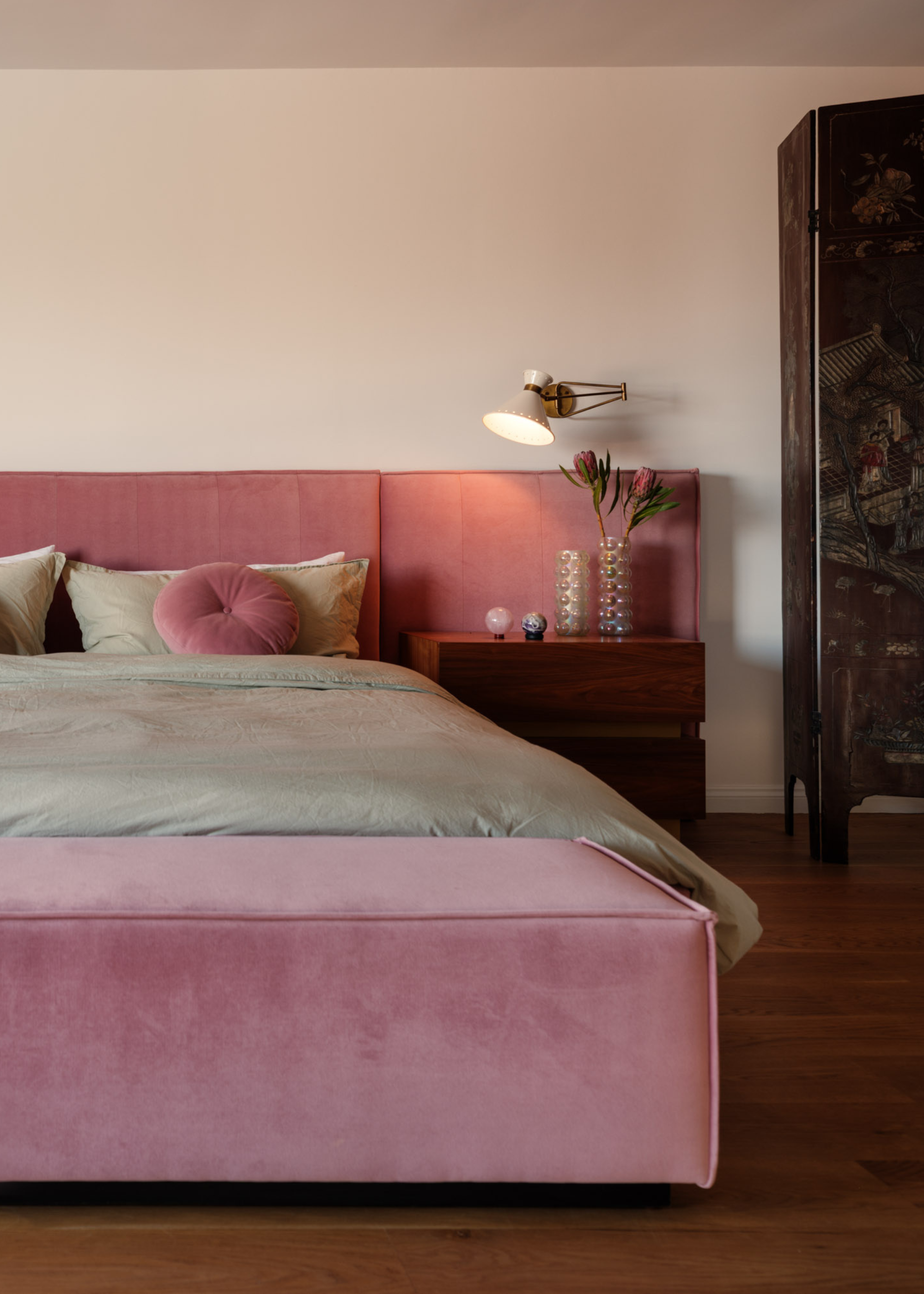 A white bedroom with a pink bed, sage green sheets, a wood sideboard with an iridescent set of vases with flowers under a wall sconce by a vintage room divider