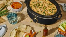 Black slow cooker full of food next to dishes of carrot and celery sticks, tortilla chips and a glass of water