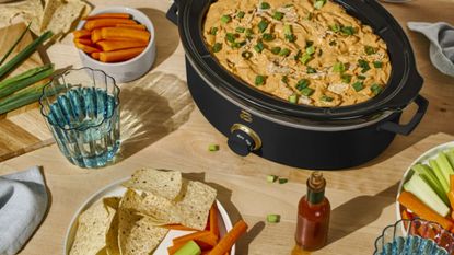 Black slow cooker full of food next to dishes of carrot and celery sticks, tortilla chips and a glass of water