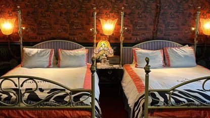 Ecclectic guest room at the Lafayette Hotel in San Diego, featuring two four-post beds with striped headboards and pink bordered bedding, along with pink toile wallpaper and a fringe antique-style table lamp.