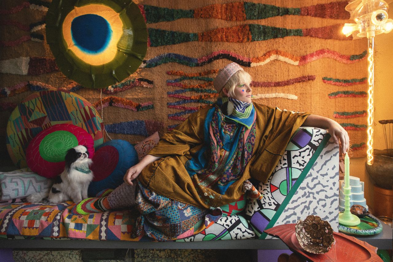 A woman dressed in vibrant, layered clothing sits on a reclining banquette with a cartoony, colorful pattern next to her dog while staring to her left. Behind her are an orangey wall tapestry and other striking home decor additions.