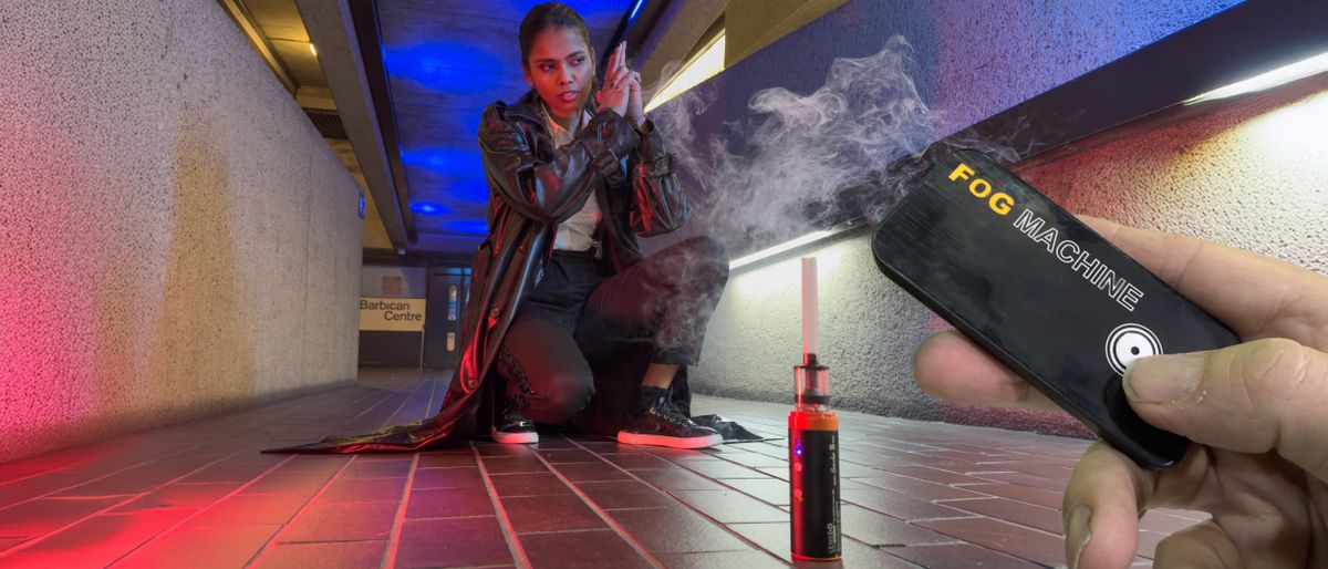 Model in a leather jacket with a gun lit by colored lights and surrounded by smoke