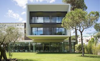 Hotel Mare e Pineta, Milano Marittima, Italy. The front of a three storey hotel with glass walls on the bottom level with a restaurant inside and a green lawn with trees on it in front of the building.