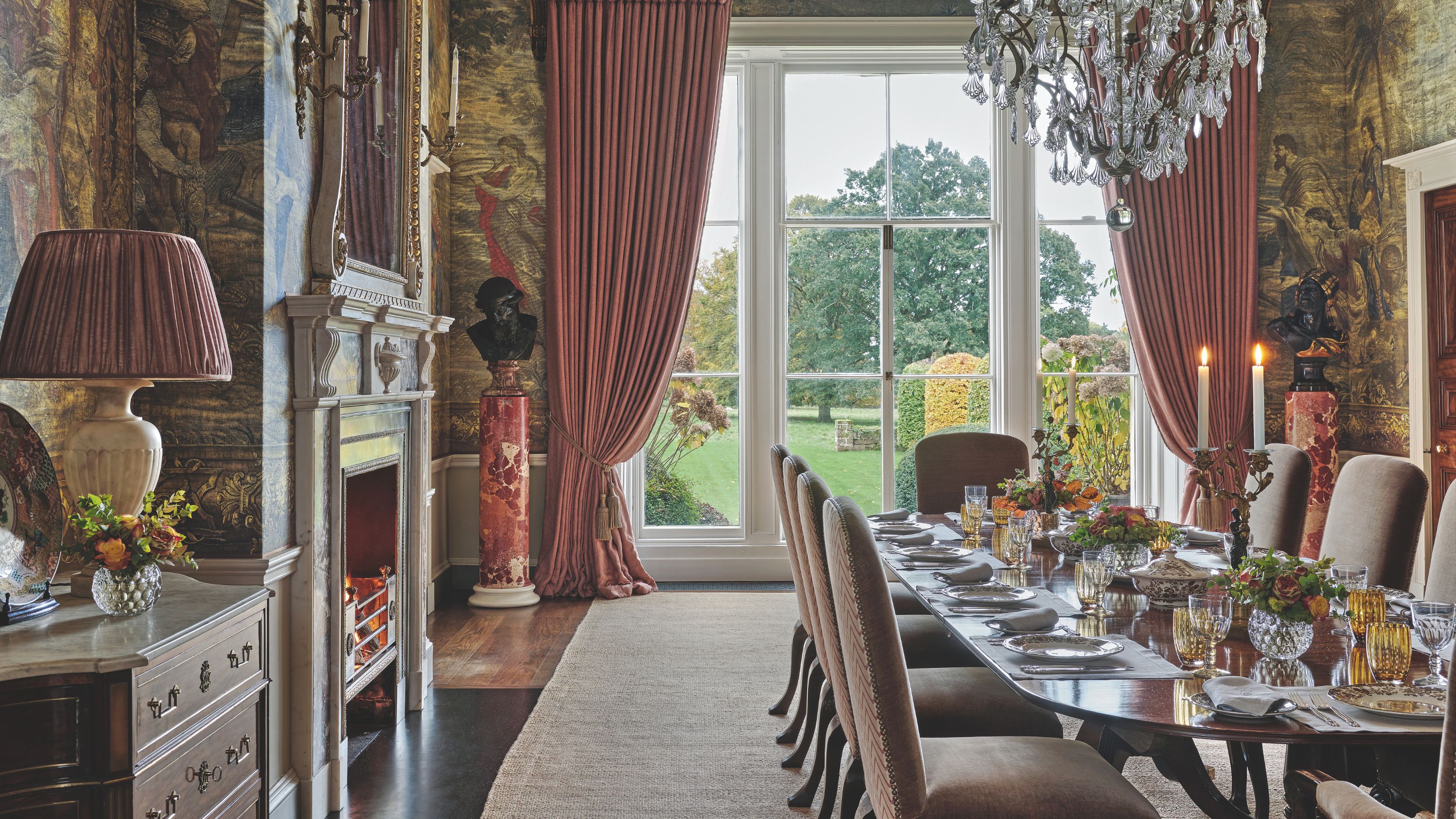 A formal dining room with expansive floor-to-ceiling windows, dramatic pink drapery, a crystal chandelier, and walls adorned with large-scale classical tapestries
