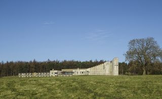 Daytime landscape image, grass lawn to the front, large stone building in the distance, forest & tall trees in the backdrop, clear blue sky