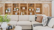 Living room space with cream L-shaped sofa and coffee table with potted flowers and books, pictured in front of floor-to-ceiling wooden shelves with miscellaneous ornaments and books.