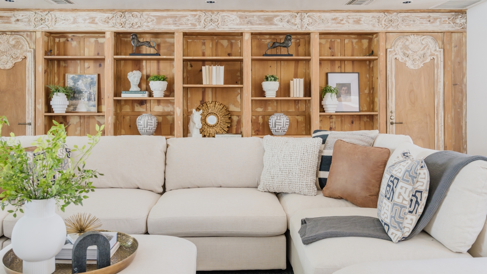 Living room space with cream L-shaped sofa and coffee table with potted flowers and books, pictured in front of floor-to-ceiling wooden shelves with miscellaneous ornaments and books.