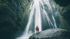 A hiker stands at the base of a waterfall and looks up at it.