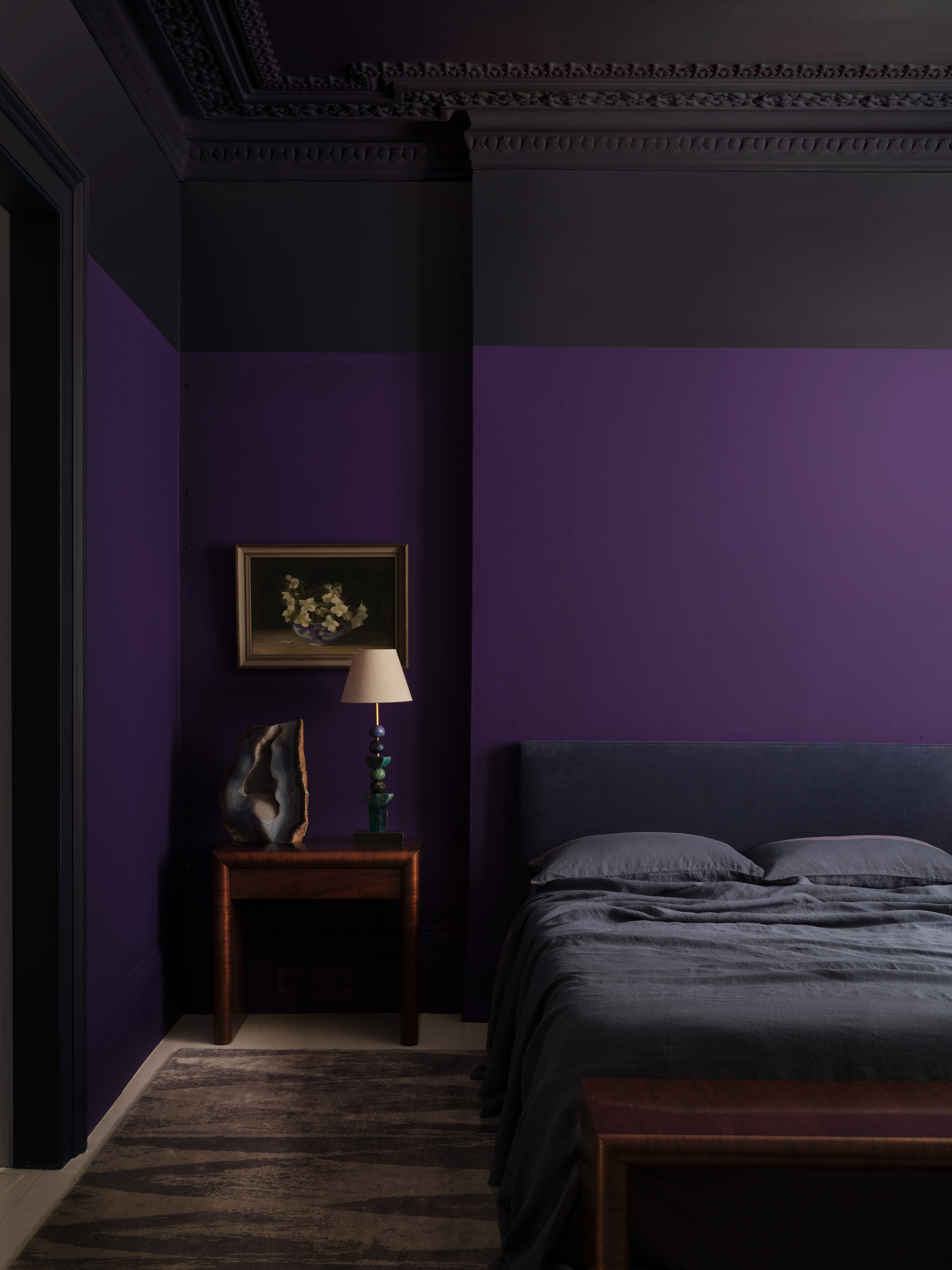 purple bedroom with black ceiling, gray sheets and black bedhead, and timber table with art