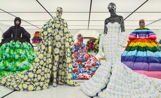 View of five models wearing floral and rainbow Moncler pieces in a room with a black and white grid style ceiling and flooring. The faces of two models can be seen and the other three models have their faces completely covered by the pieces. Two of the models with their faces covered are also wearing white sunglasses