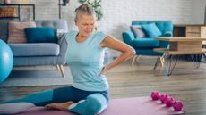 A woman sits on a yoga mat in a living room, with light dumbbells behind her. One leg is outstretched and the other is bent on the floor. Both her hands are on her back and she is frowning. Behind her is a couch, chair, coffee table and plant.