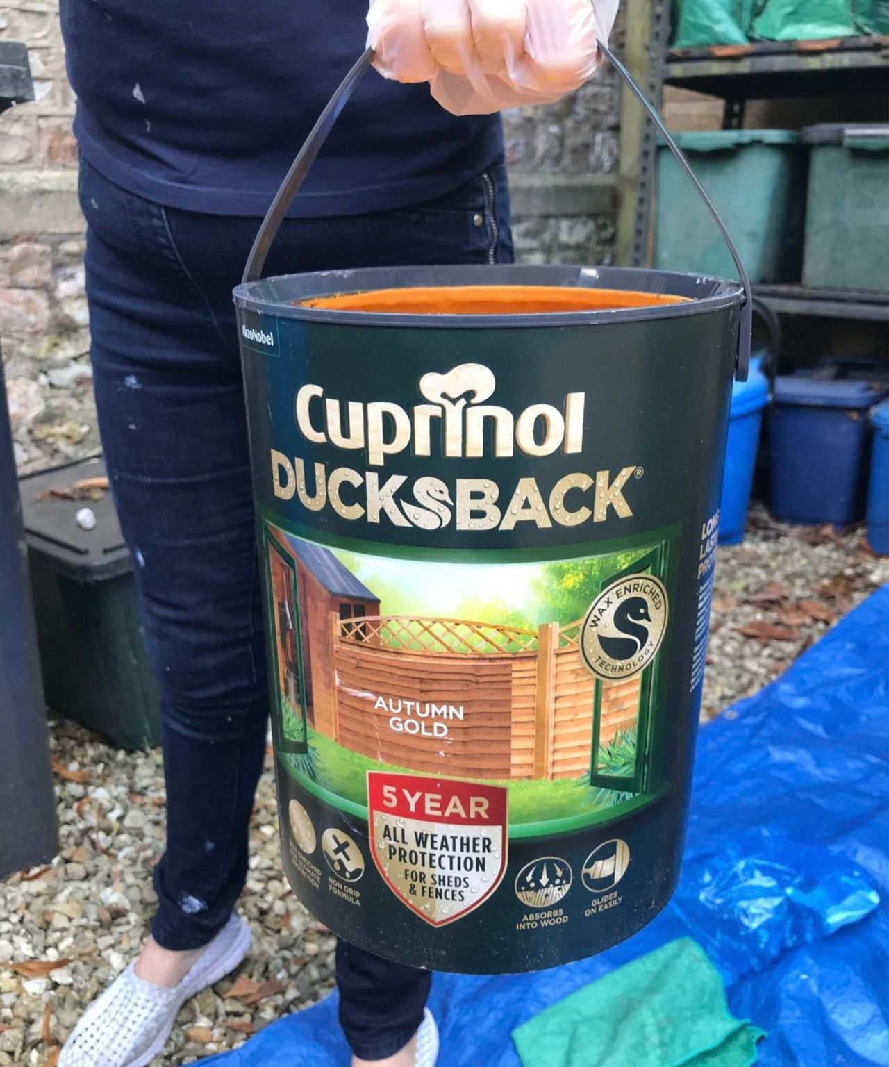 Person holding a tin of Cuprinol Ducksback wood treatment in Autumn Gold, with gloved hand on the handle.