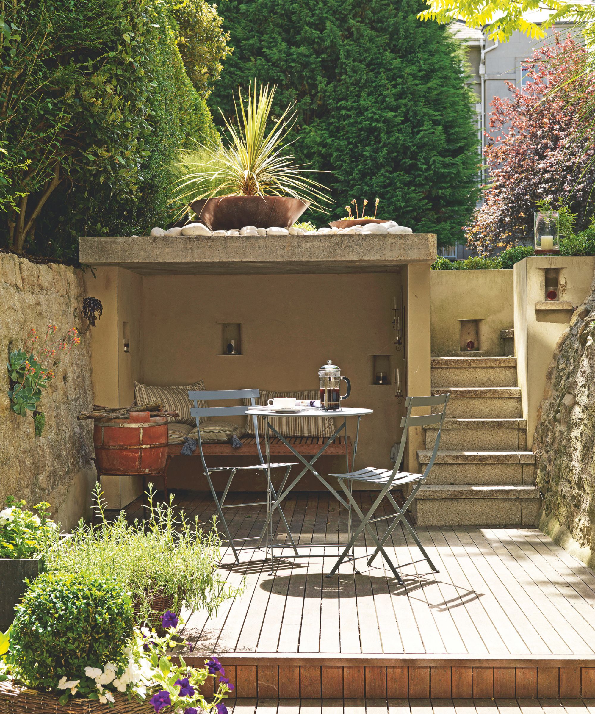 Small patio with stairs leading up to the garden, a level with a wood panelled floor, a metal bistro set, and an alcove with a built-in bench and striped cushions.