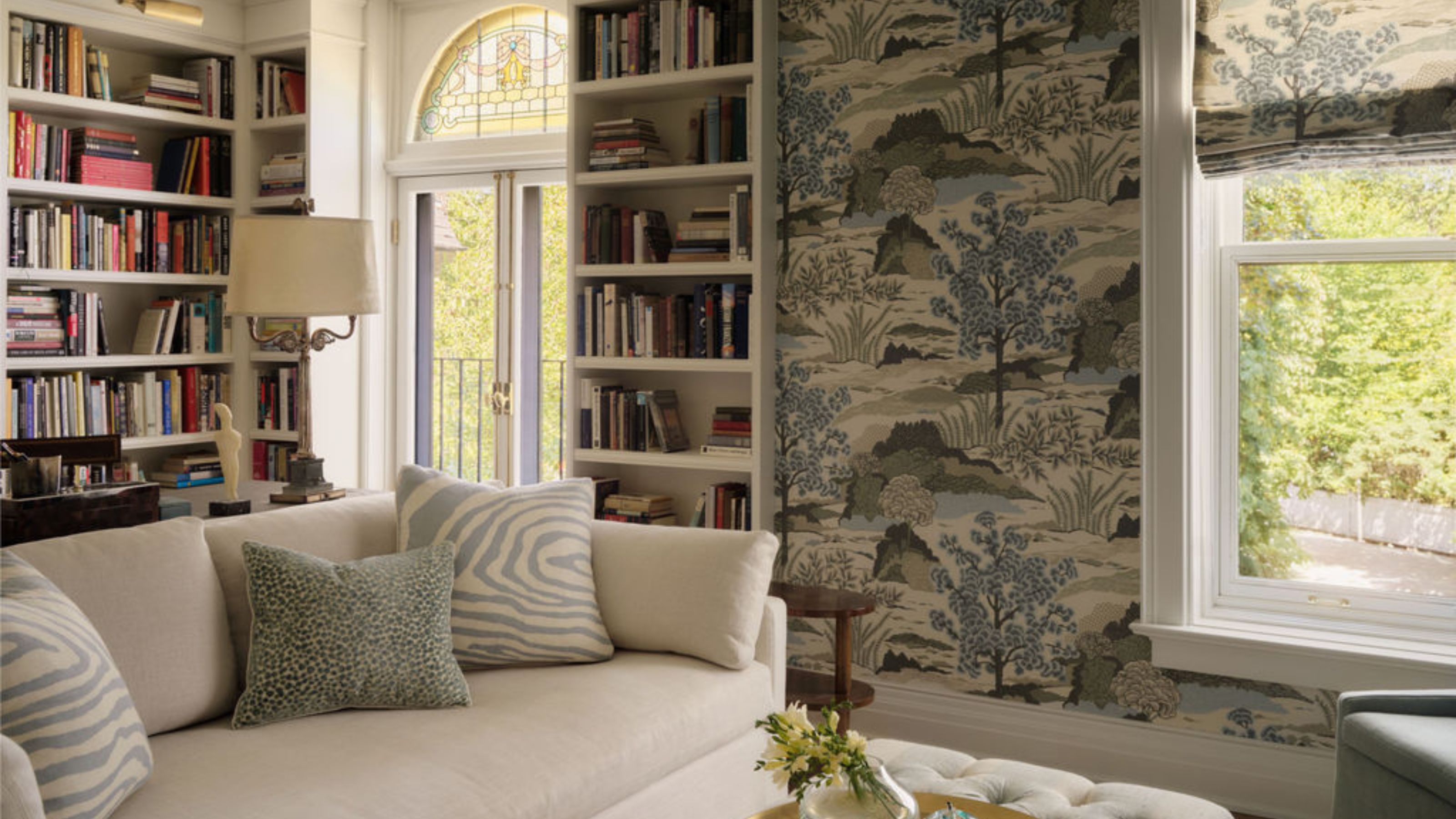 a layered home library with stacked book shelves, stain glass window, printed wallpaper and a large white couch