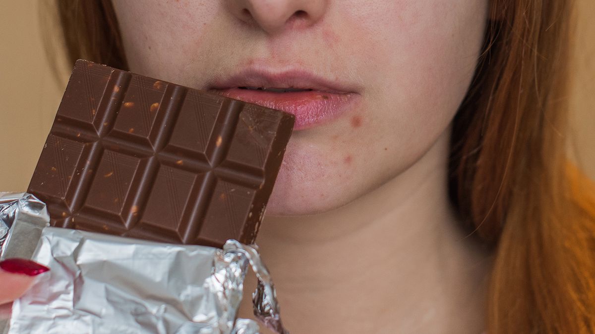 close up of young woman with pimples around her mouth holding up a chocolate bar as if to eat it