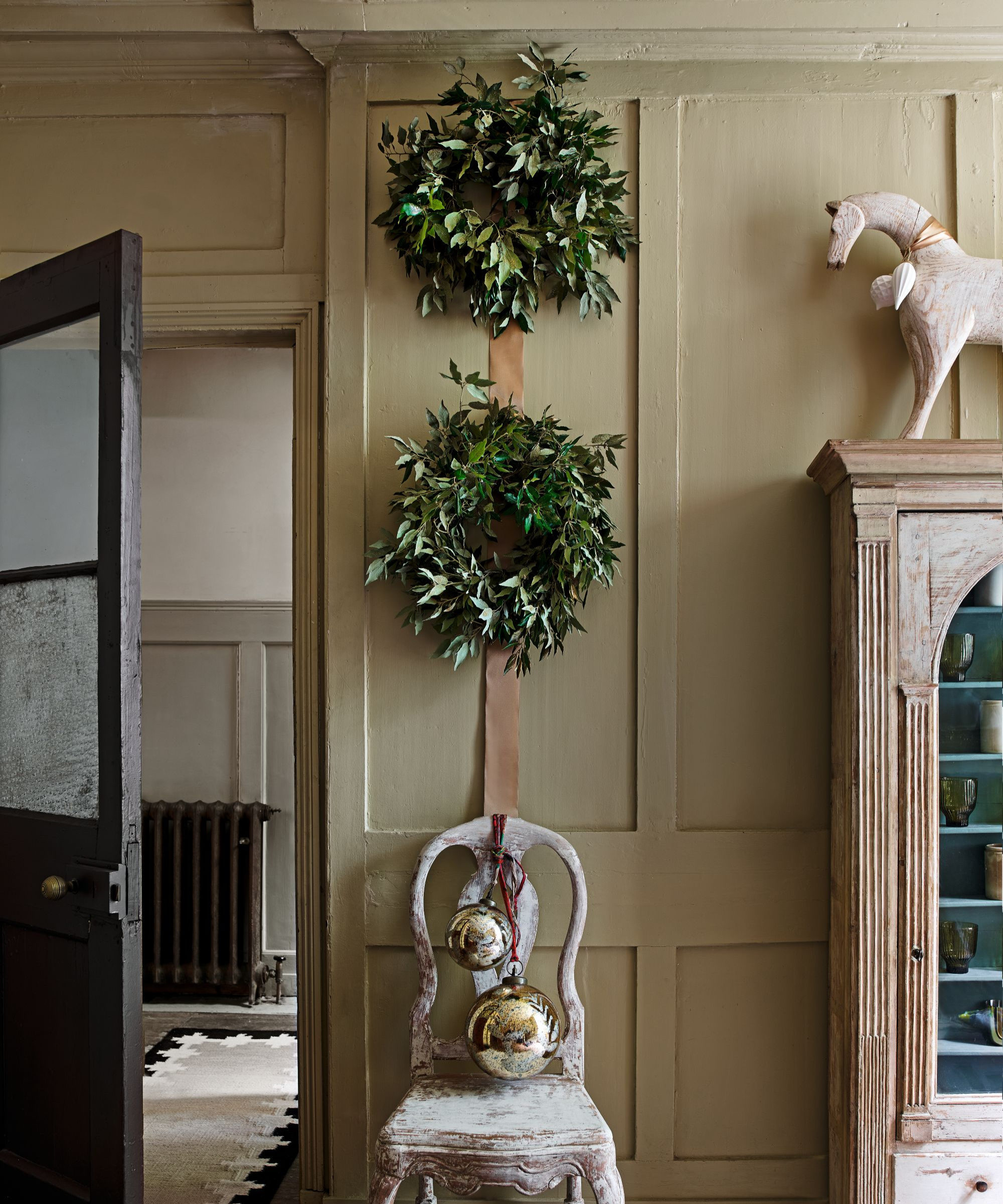 a set of two wreaths hanging on a wall, one above the other. A rustic chair below with two large ornaments hanging from it