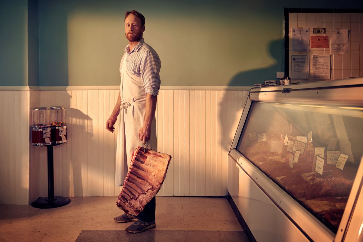 A butcher in an apron stands holding a large slab of meat in a shop, with a glass display case and gum ball machines in the background