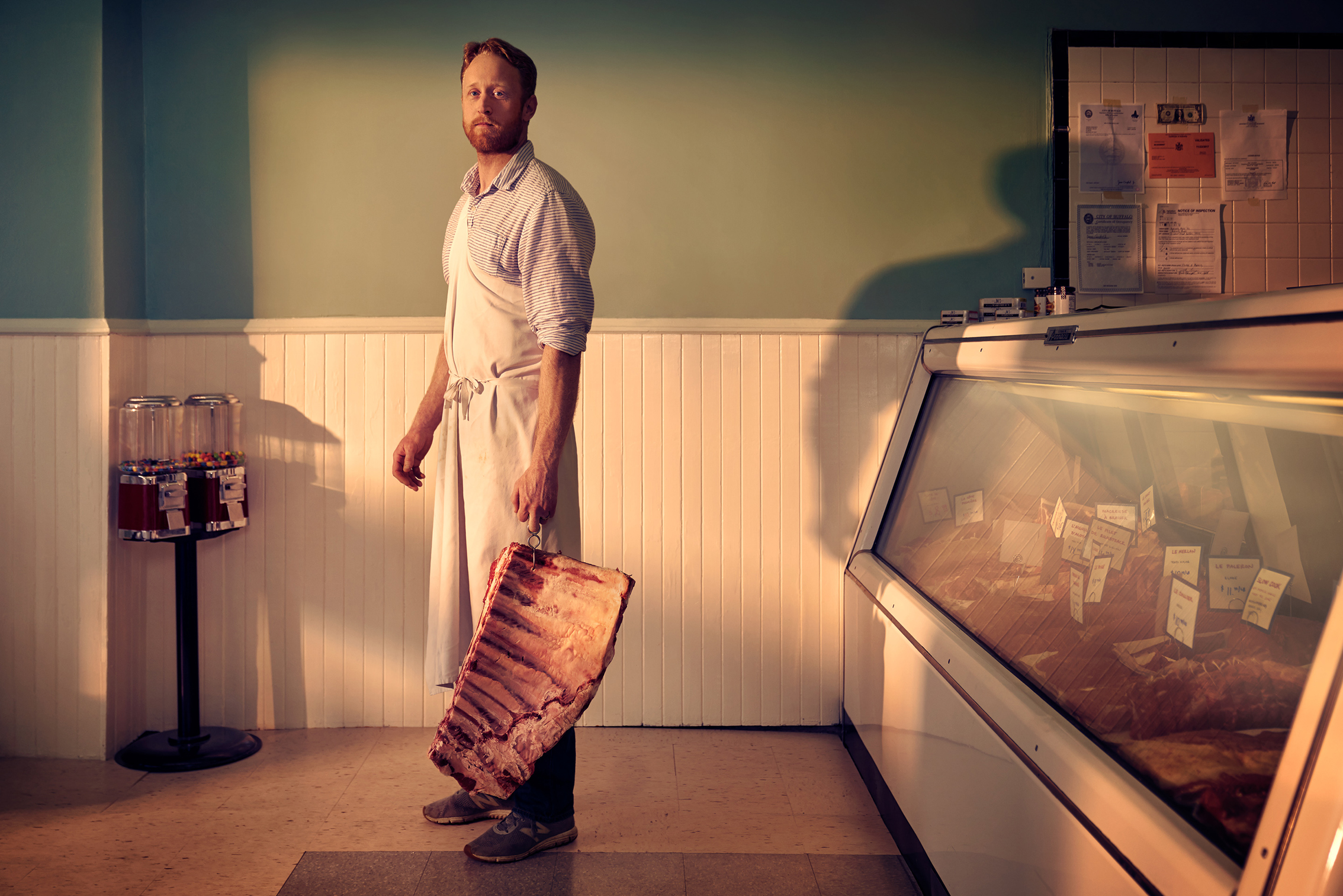 A butcher in an apron stands holding a large slab of meat in a shop, with a glass display case and gum ball machines in the background