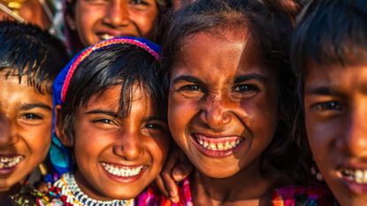 Close-up image of a group of four Indian children (with the face of another more blurred behind them) who are smiling at the camera. The sun in shining on their faces