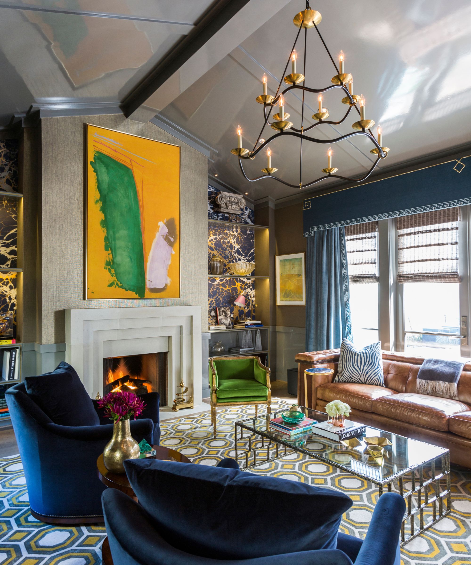 A formal living room with a high sloped ceiling painted light gray, in a high-gloss, mirror-like finish, Dark blue armchairs and window treatments, a bright yellow and green artwork above the fireplace, and a geometric print rug.