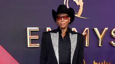 RuPaul wearing a black cowboy hat and jacket, standing in front of a purple wall that says 'Emmys' in gold writing behind it