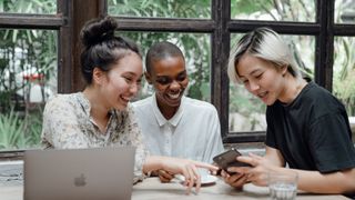 Three smiling women sat around desk, as one shows them her smartphone screen