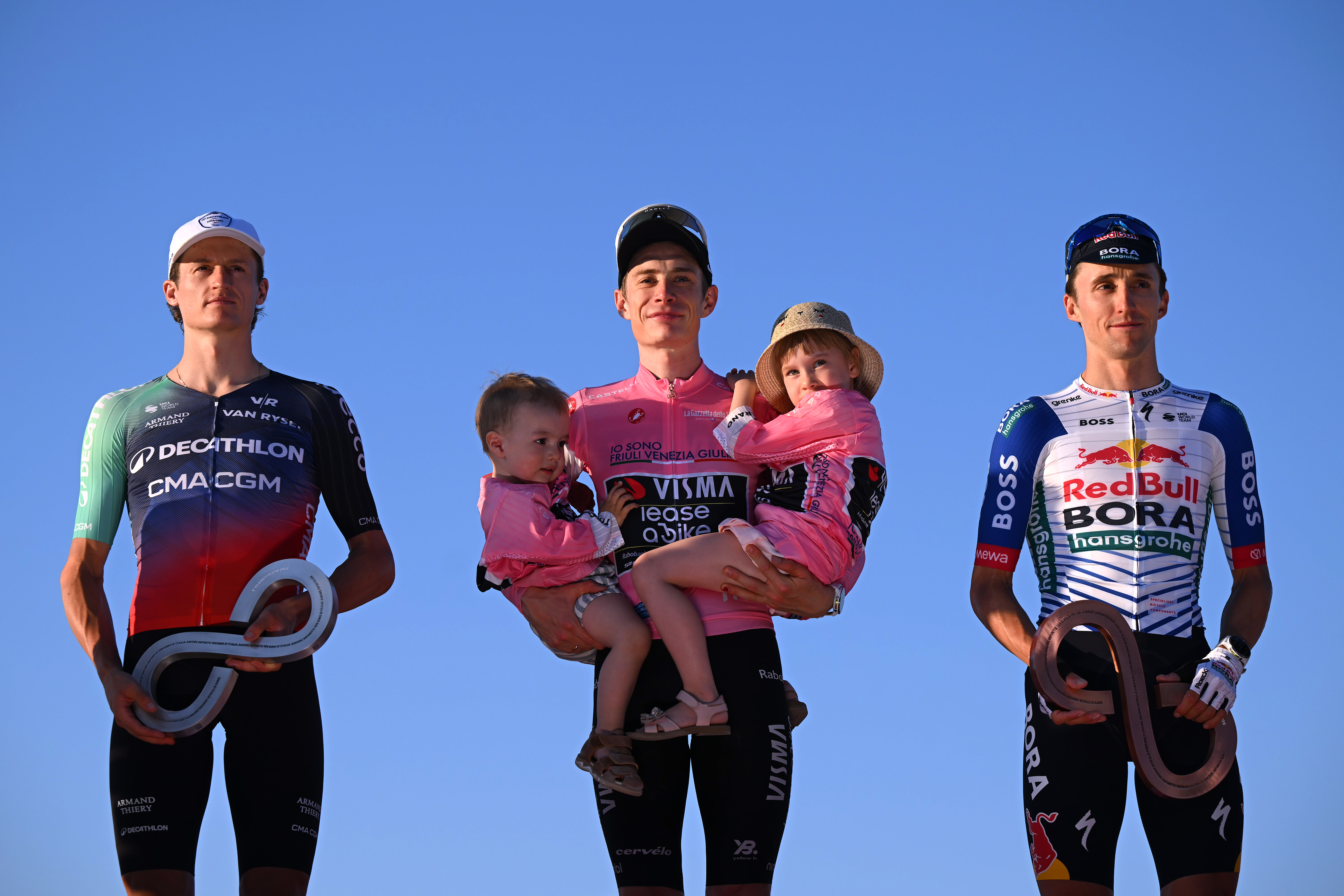 Gall, Vingegaard holding his children, and Hindley on the podium against a blue sky