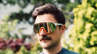 A white man with a moustache wears blue and orange sunglasses