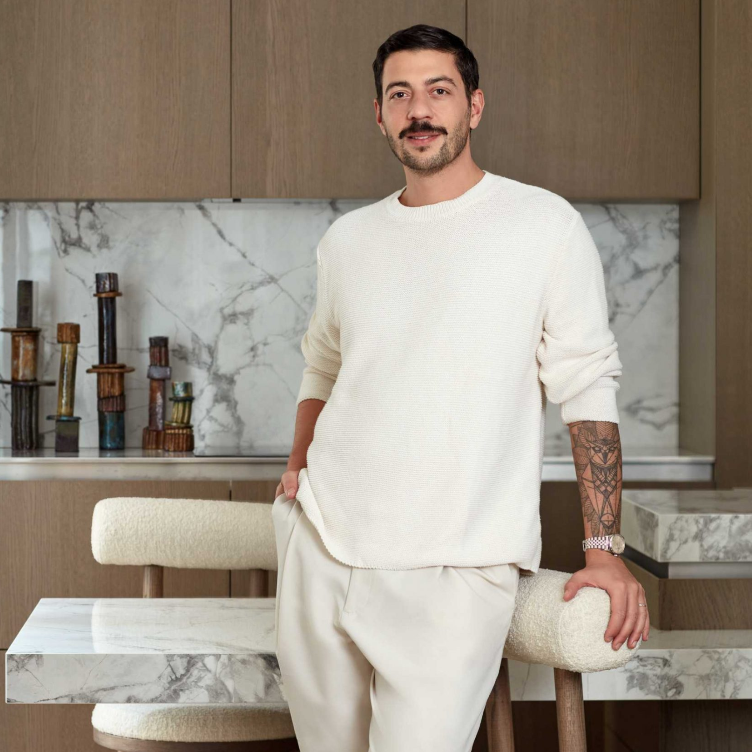 A man is pictured in a white round neck sweater and trousers leaning against a boucle chair in a kitchen