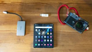 Folding phone, leica camera and dongle and small card reader layout on top of a wooden table